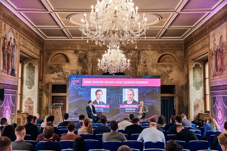 Conference audience facing a stage and large presentation screen beneath chandeliers